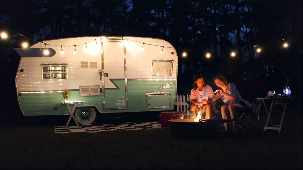 Two girls next to a retro RV camping and roasting marshmellows